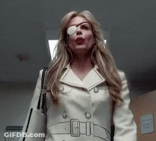 A Woman With A Bandage On Her Eye Is Wearing A White Trench Coat And Sunglasses . GIF