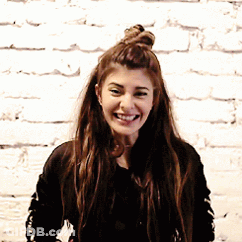 A Woman With A Bun On Her Head Is Smiling In Front Of A White Brick Wall GIF