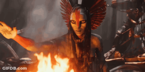 A Woman With A Feathered Headdress Is Standing In Front Of A Fire GIF