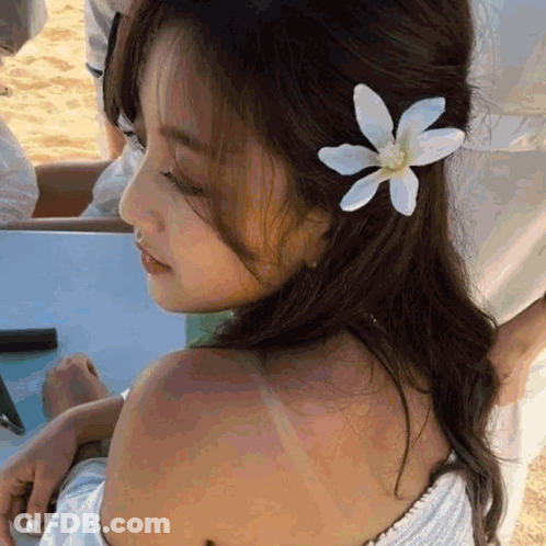 A Woman With A Flower In Her Hair Is Sitting On The Beach GIF