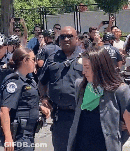 A Woman With A Green Bandana On Her Neck Is Being Escorted By Police GIF