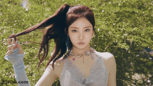 A Woman With A Ponytail And A Star Necklace Stands In Front Of Trees GIF