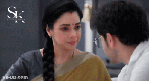 A Woman With A Red Dot On Her Forehead Looks At A Man With Glasses In Front Of A Sign That Says Sk GIF