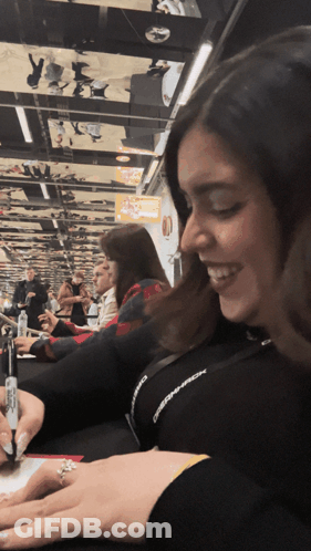 A Woman With A Ring On Her Finger Is Smiling In Front Of A Crowd Of People GIF
