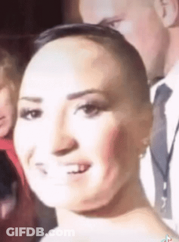 A Woman With A Shaved Head Is Smiling In Front Of A Man In A Suit And Tie . GIF