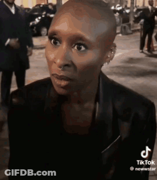 A Woman With A Shaved Head Is Wearing A Black Jacket And Earrings And Looking At The Camera . GIF