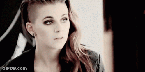 A Woman With A Shaved Head Is Wearing Earrings And A Black Jacket . GIF