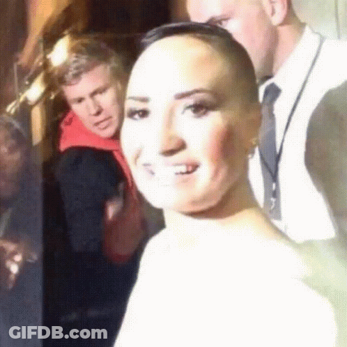 A Woman With A Shaved Head Smiles In Front Of A Man In A Suit GIF
