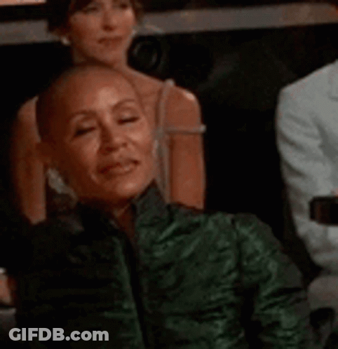A Woman With A Shaved Head Wearing A Green Jacket Is Sitting In A Theatre . GIF