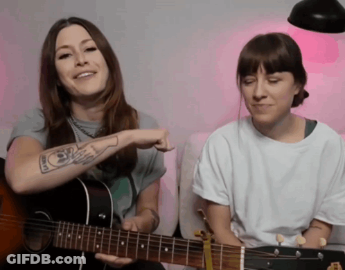 A Woman With A Tattoo On Her Arm Is Playing A Guitar Next To Another Woman GIF