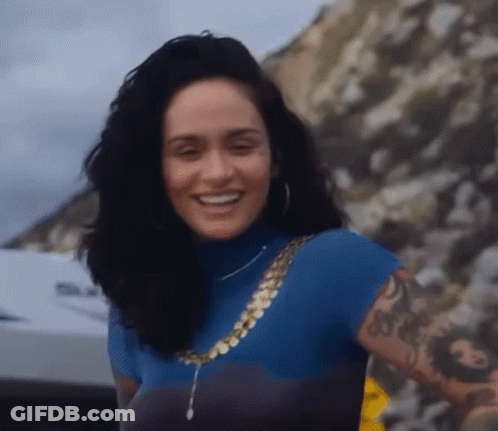 A Woman With A Tattoo On Her Arm Is Smiling While Wearing A Blue Turtleneck And A Gold Chain . GIF