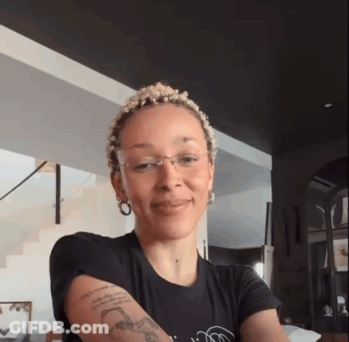 A Woman With A Tattoo On Her Arm Is Wearing Glasses And A Black Shirt GIF