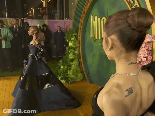 A Woman With A Tattoo On Her Back Stands In Front Of A Green Door That Says Wizard Of Oz GIF