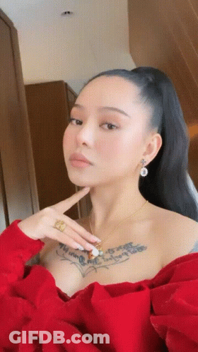 A Woman With A Tattoo On Her Chest Is Wearing A Red Top And Earrings GIF