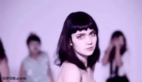 A Woman With Black Hair Is Standing In Front Of A Group Of People And Looking At The Camera . GIF