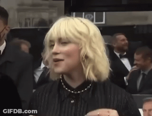 A Woman With Blonde Hair And A Black Shirt Is Standing In A Crowd . GIF