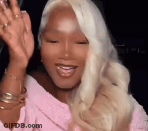 A Woman With Blonde Hair And A Pink Shirt Is Waving At The Camera . GIF