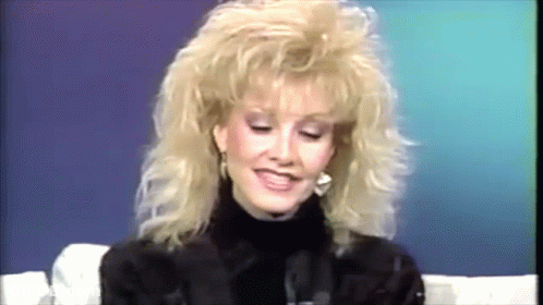 A Woman With Blonde Hair And A Turtleneck Is Smiling GIF