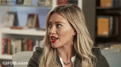 A Woman With Blonde Hair And Red Lips Is Sitting In Front Of A Bookshelf And Making A Funny Face . GIF