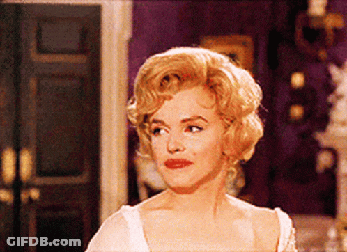 A Woman With Blonde Hair And Red Lips Stands In A Room GIF