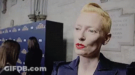A Woman With Blonde Hair And Red Lipstick Is Standing In Front Of A Sign That Says ' Ladies Of The Race ' . GIF