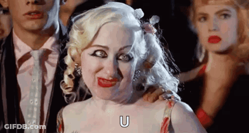 A Woman With Blonde Hair Is Making A Funny Face While Standing Next To A Man In A Suit . GIF