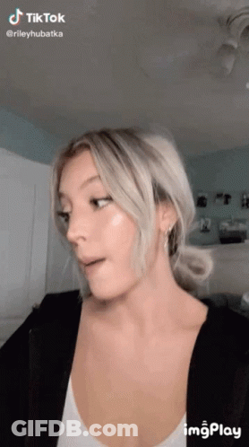 A Woman With Blonde Hair Is Making A Funny Face While Wearing A White Tank Top And A Black Jacket . GIF