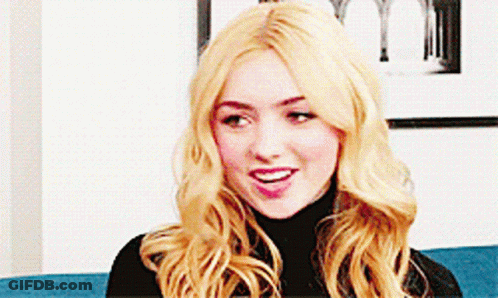A Woman With Blonde Hair Is Sitting On A Blue Couch Wearing A Black Turtleneck . GIF