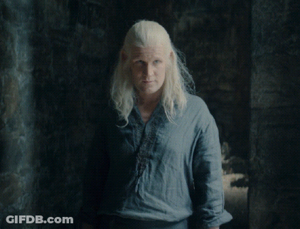 A Woman With Blonde Hair Wearing A Blue Shirt Stands In A Dark Room GIF