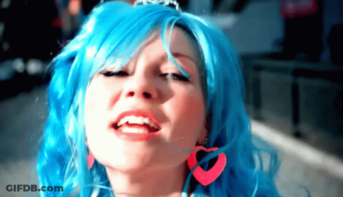 A Woman With Blue Hair And Pink Heart Shaped Earrings Smiles GIF