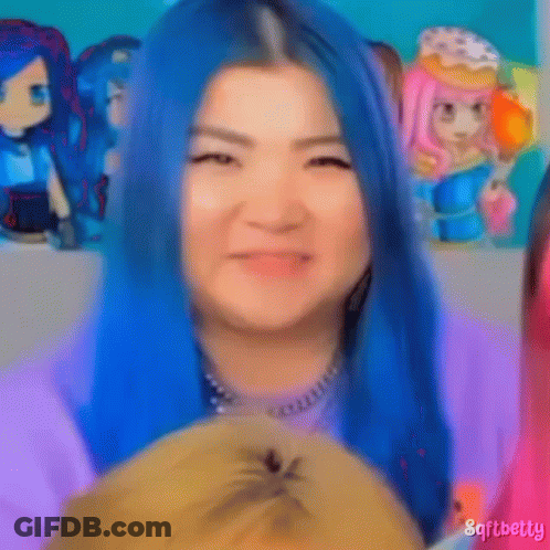 A Woman With Blue Hair Is Holding A Stuffed Animal In Front Of A Group Of Dolls . GIF