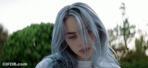 A Woman With Blue Hair Is Standing In Front Of A Bush With Her Eyes Closed And Her Hair Blowing In The Wind . GIF