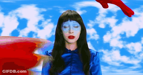 A Woman With Blue Paint On Her Face Stands In Front Of A Blue Sky With Clouds GIF
