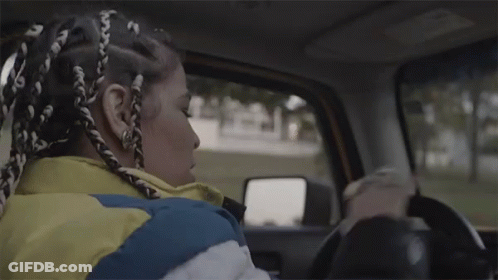 A Woman With Braids Is Driving A Car While Wearing A Yellow Jacket . GIF
