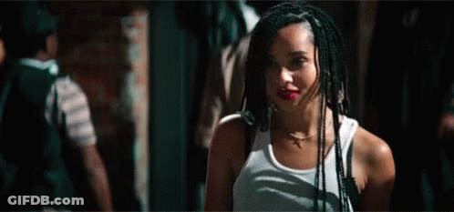 A Woman With Braids Is Wearing A White Tank Top And Standing In A Dark Room . GIF