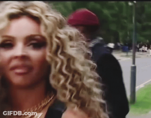 A Woman With Curly Blonde Hair Is Standing Next To A Man On A Sidewalk . GIF