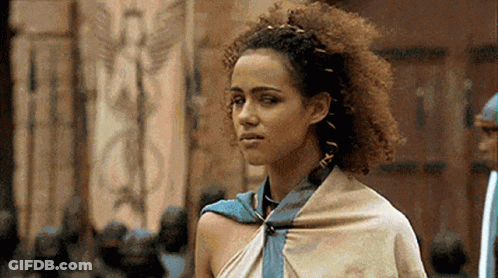 A Woman With Curly Hair And A Scarf Around Her Neck Is Standing In Front Of A Building . GIF