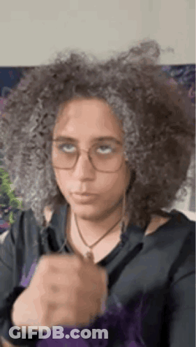 A Woman With Curly Hair And Glasses Is Wearing A Black Shirt And A Purple Jacket . GIF