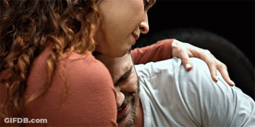 A Woman With Curly Hair Is Hugging A Man With His Eyes Closed GIF