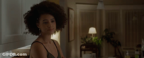 A Woman With Curly Hair Is Sitting At A Table In A Dark Room GIF