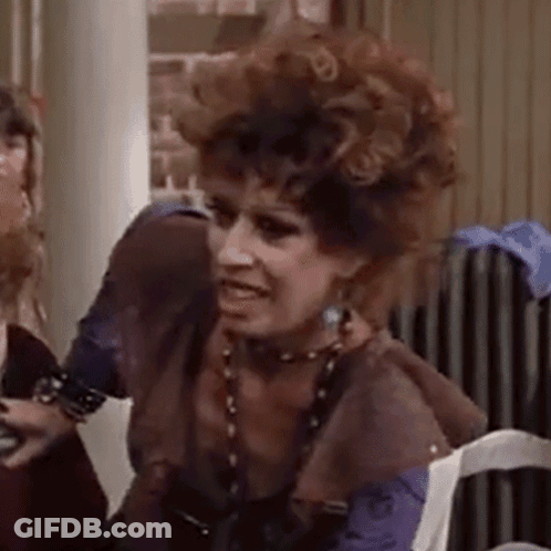 A Woman With Curly Hair Is Sitting In A Chair And Making A Face . GIF