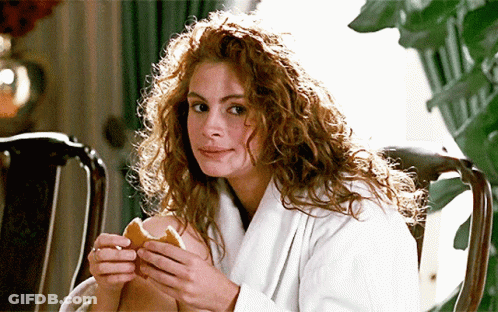 A Woman With Curly Hair Is Sitting In A Chair Holding A Sandwich GIF