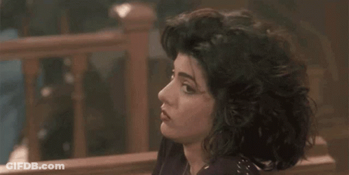 A Woman With Curly Hair Is Sitting On A Bench In Front Of A Wooden Railing . GIF