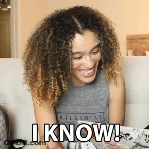 A Woman With Curly Hair Is Sitting On A Couch And Smiling While Holding A Magazine And Says I Know GIF