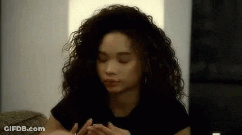 A Woman With Curly Hair Is Sitting On A Couch With Her Eyes Closed And Her Hands On Her Face . GIF