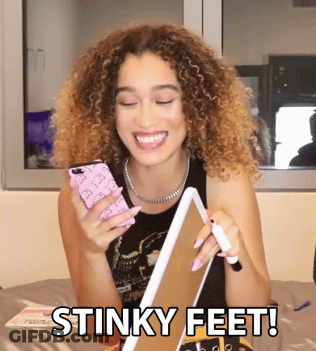 A Woman With Curly Hair Is Smiling While Holding A Cell Phone And A Piece Of Paper With The Words Stinky Feet Below Her GIF