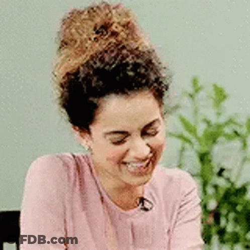 A Woman With Curly Hair Is Smiling While Wearing A Pink Shirt And Earrings . GIF