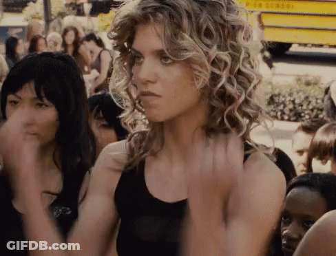 A Woman With Curly Hair Is Standing In Front Of A Crowd Of People And A Yellow School Bus GIF