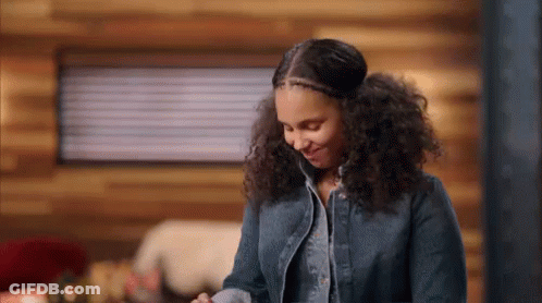 A Woman With Curly Hair Is Standing In Front Of A Wooden Wall In A Cabin And Smiling . GIF