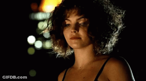 A Woman With Curly Hair Is Wearing A Black Bra And Looking At The Camera In The Dark . GIF